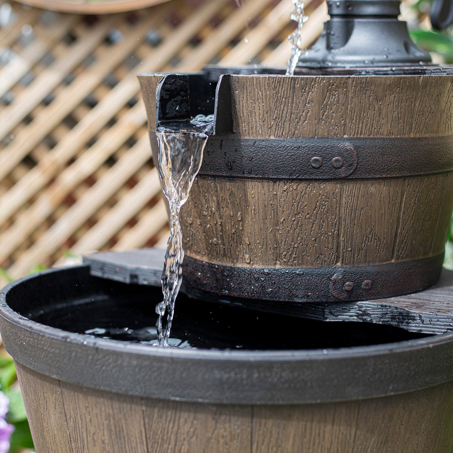 A close-up view of La Hacienda's Whiskey Bowls, a rustic self-contained fountain crafted from weathered wooden barrels, showcases water cascading elegantly down. This charming outdoor water feature is perfectly positioned against a lattice backdrop and surrounded by small plants. The barrels' metal bands enhance the piece's allure, echoing the charm of its namesake.