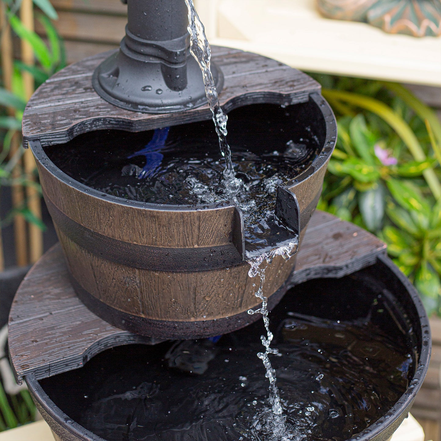 This striking outdoor water feature, the Whiskey Bowls by La Hacienda, is a self-contained fountain with a rustic wooden barrel design. Water cascades elegantly from a black spout into the top basin and down to the lower one, while surrounding greenery enhances its natural charm.