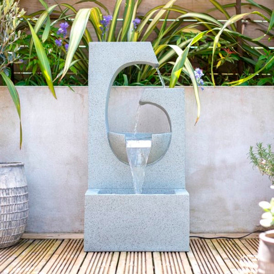 The Ango Falls by La Hacienda creates a stunning contemporary garden statement with its geometric design, where water gracefully cascades from a curved cutout into a basin. This modern outdoor water fountain is beautifully complemented by wooden decking and lush green plants with purple flowers.