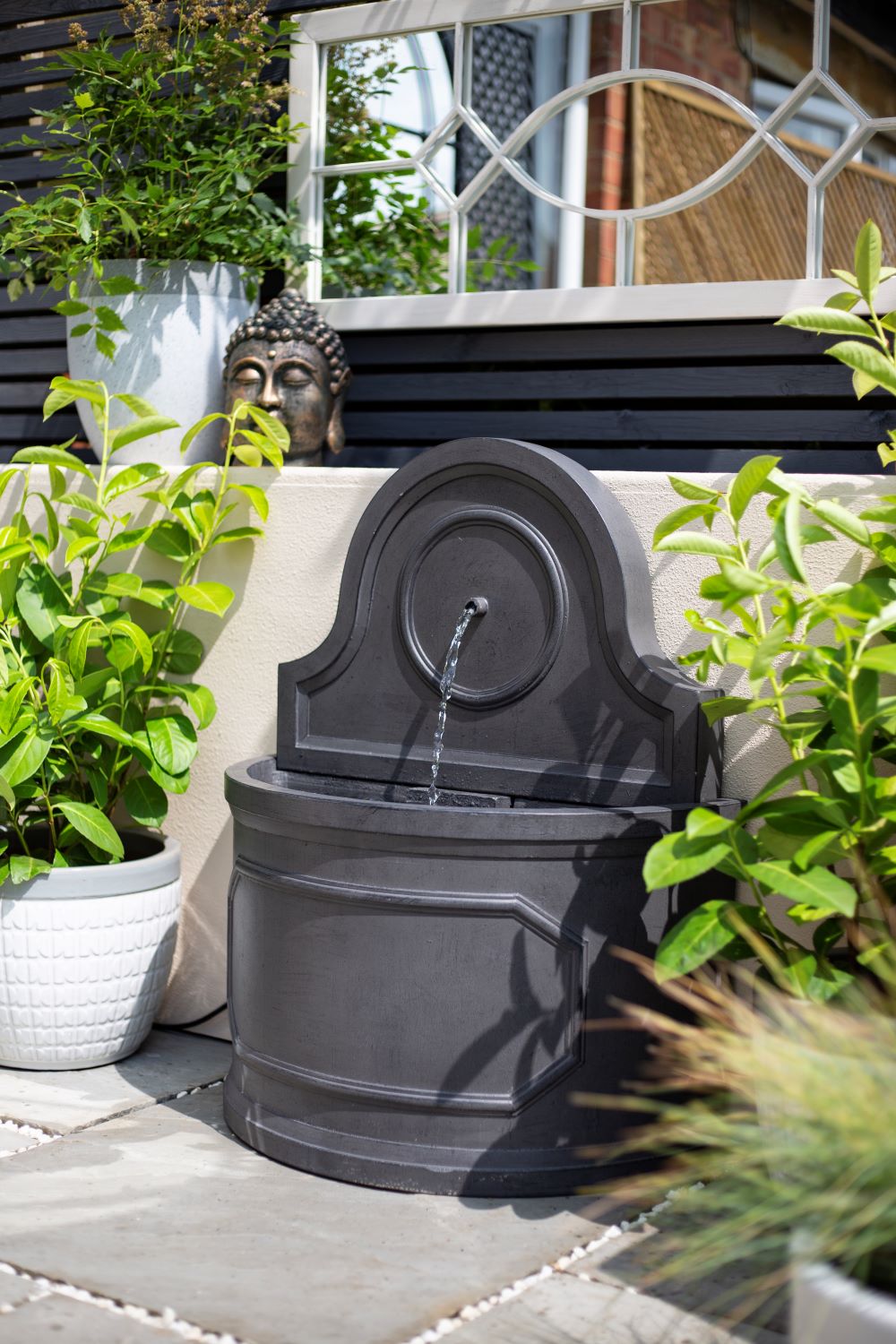 The La Hacienda Winchcombe Springs Water Feature, a decorative dark gray stone fountain, creates a serene garden ambiance on a patio surrounded by lush green plants in white pots. A stone Buddha head nestles among the plants, and a large mirror with a geometric pattern hangs on the wall behind this contemporary La Hacienda water feature.