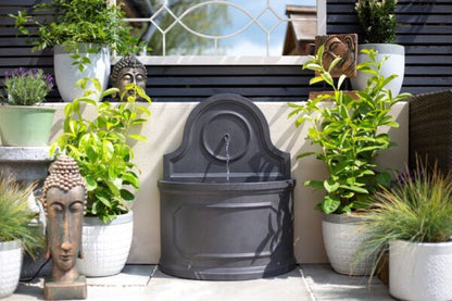 The La Hacienda Winchcombe Springs Water Feature is surrounded by lush, green plants in white pots. Two Buddha head sculptures and a Buddha wall art piece are placed among the foliage, enhancing the garden ambiance. A lattice fence and the blue sky are visible in the background.