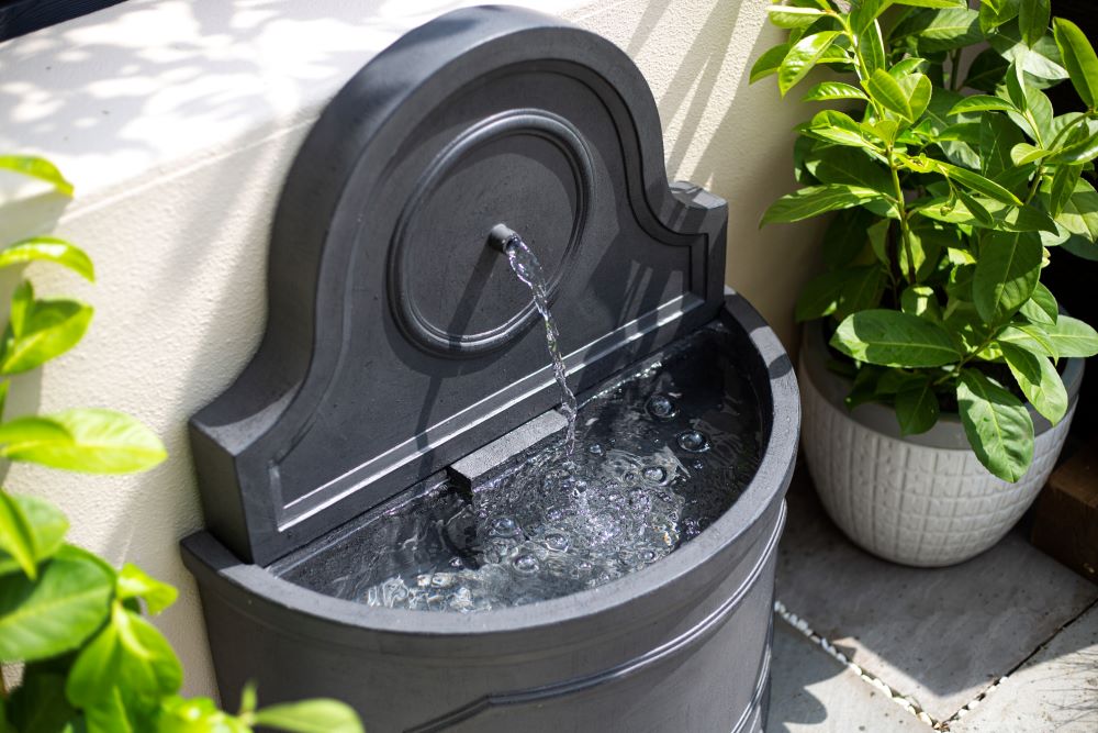 The La Hacienda Winchcombe Springs Water Feature, a small, gray stone wall fountain with a classic arched design, is depicted with water gently flowing from a spout into a semi-circular basin below. This elegant piece by La Hacienda is flanked by potted green plants and set against a light-colored wall in an outdoor setting, enhancing the garden ambiance.