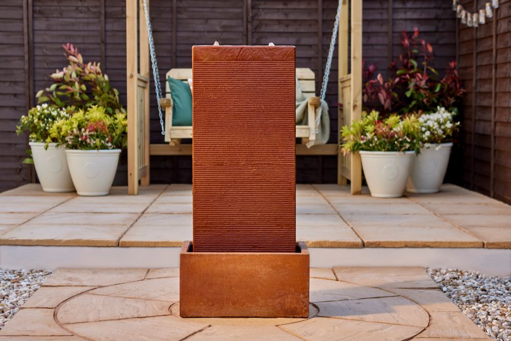 A rectangular, rust-colored La Hacienda Amber Falls Water Feature stands in an outdoor patio area, embodying the elegant design of a garden fountain. Behind it, there is a wooden swing adorned with teal and beige cushions, surrounded by potted plants with vibrant green, yellow, and red foliage. The setting has a tranquil and neatly arranged look.