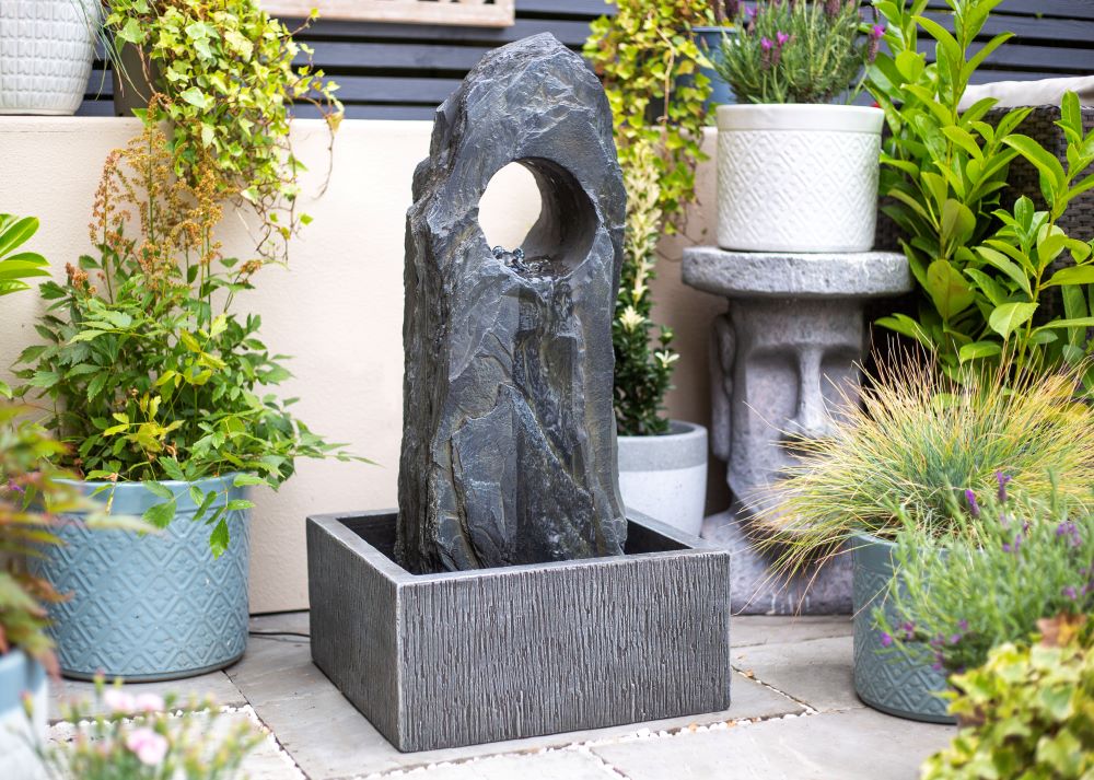 A serene garden scene featuring the La Hacienda Cambrian Monolith Water Feature, a rectangular stone fountain with a tall, dark slate rock that has a circular hole near the top. This solar-powered masterpiece from La Hacienda is surrounded by various potted plants, including leafy greens and colorful flowers, enhancing the lush and vibrant garden ambiance.