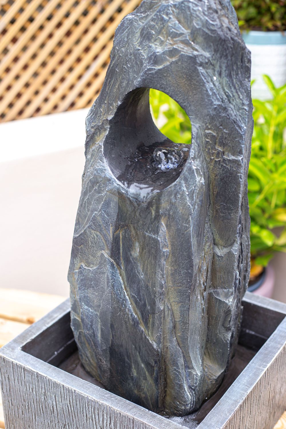 The La Hacienda Cambrian Monolith is a tall, textured water fountain with a natural appearance, featuring a circular opening at the top and standing on a square base. Water gently flows through the opening, with a blurred background of lattice fencing and green plants.