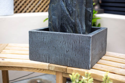 A close-up of La Hacienda's Cambrian Monolith reveals a textured, rectangular stone water feature positioned on a wooden bench. The fountain showcases a dark, striated surface, set against a cream wall with greenery in the background.