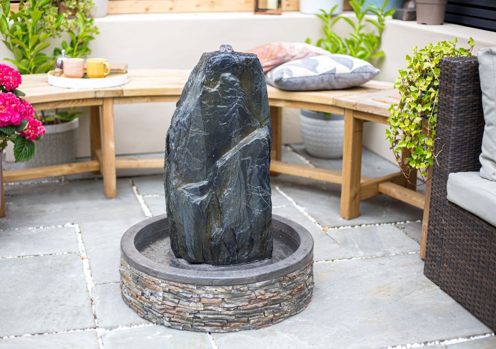 A serene garden patio is centered around a tall, elegant water fountain from La Hacienda, known as the Snowdonia Monolith, resting in a circular basin. Wooden benches with cushions encircle it, accompanied by green plants and a blossoming pink hydrangea bush, crafting a peaceful and inviting atmosphere.