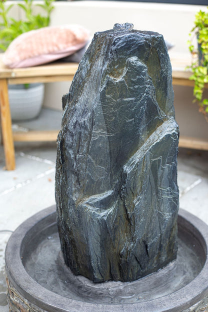 A tall, natural stone water fountain from La Hacienda, known as the Snowdonia Monolith, stands majestically in a circular basin on the patio. Water delicately trickles down its textured surface while plants and outdoor furniture create a cozy backdrop.