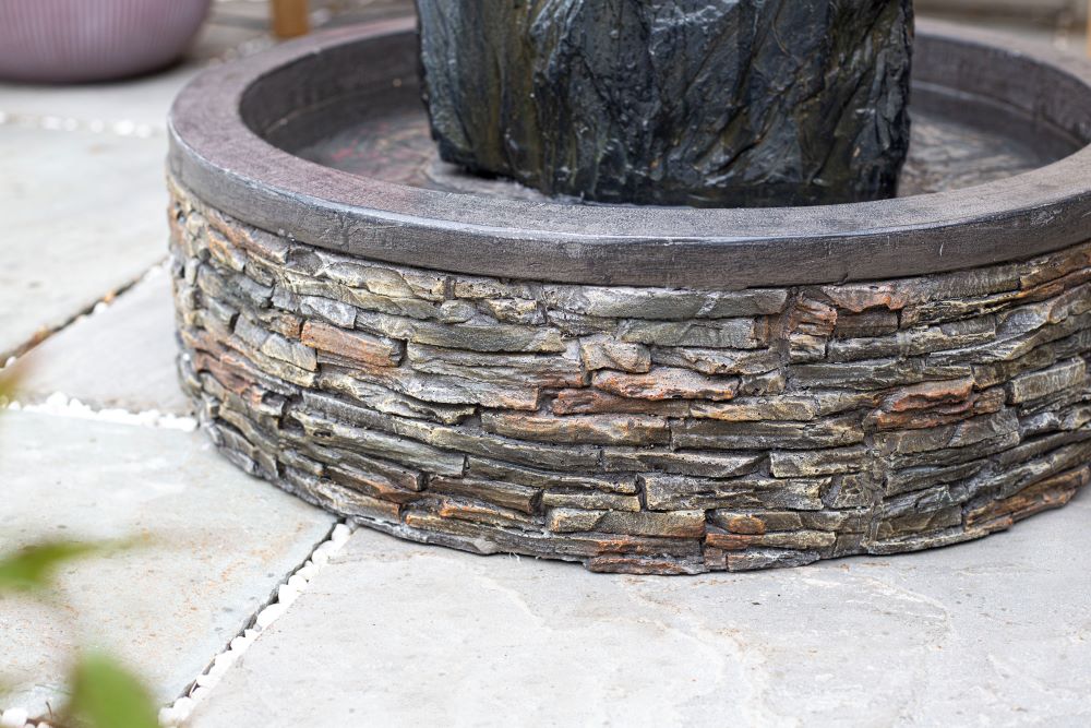 Close-up shot of the La Hacienda Snowdonia Monolith Water Feature in a garden. The base of this La Hacienda water feature is made of stacked, uneven stones. The surrounding surface features large, light-colored paving stones. The focus highlights the texture and detail of the stonework.