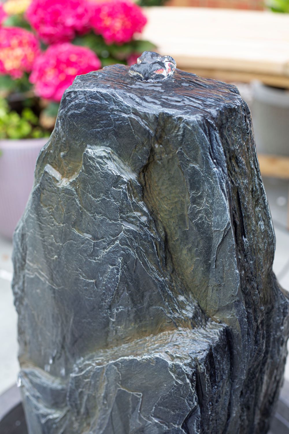 A close-up view of the La Hacienda Snowdonia Monolith Water Feature with water trickling down its textured surface. In the background, vibrant pink flowers and wooden benches enhance the garden ambiance, creating a colorful and serene setting. This self-contained water feature by La Hacienda adds an elegant touch to your outdoor space.