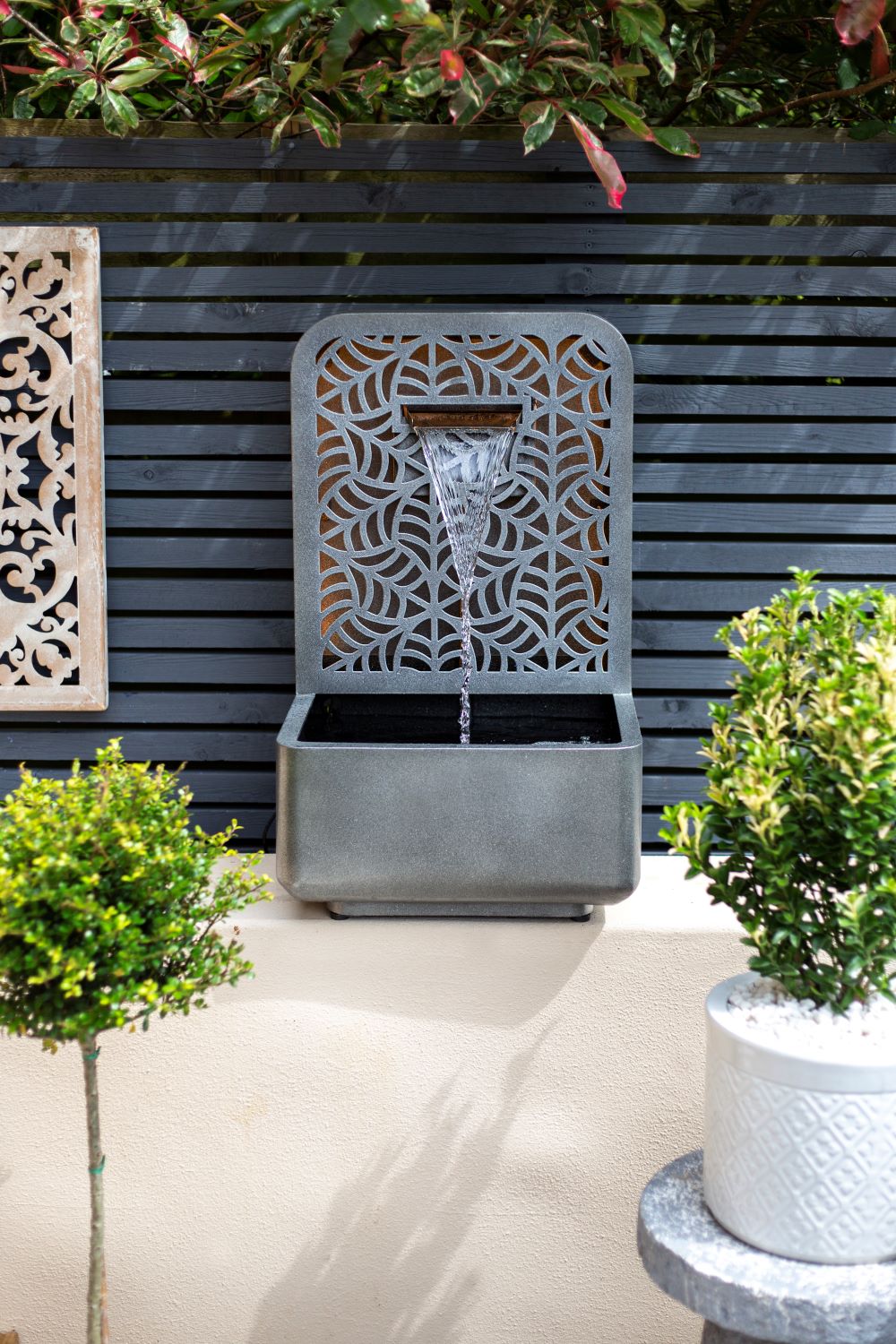 A La Hacienda Rene Falls Water Feature by La Hacienda, featuring intricate patterns, is mounted on a black slatted fence. Water gracefully cascades from the top into a square basin below, creating a tranquil outdoor setting with potted green plants and a stone wall.