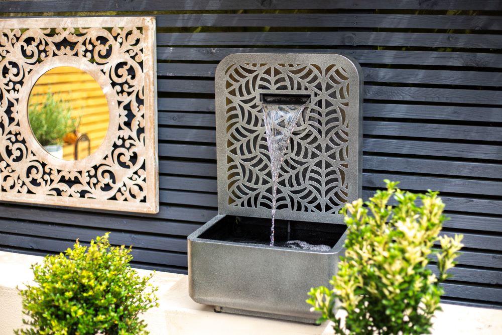 The RENÉ FALLS by La Hacienda features an ornate art deco style, with water cascading into a rectangular basin set against a dark horizontal slat wall. Nearby, a round decorative mirror enhances the aesthetic. This self-contained water feature is beautifully complemented by lush green plants in the foreground.