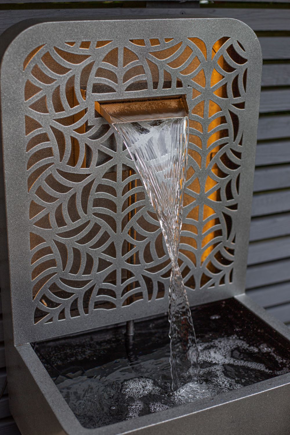 The La Hacienda Rene Falls Water Feature by La Hacienda showcases exquisite geometric patterns on steel, with water cascading smoothly from a horizontal spout into a rectangular basin below. Gentle yellow lighting behind the metal designs creates a warm glow, enhancing the serene outdoor atmosphere.