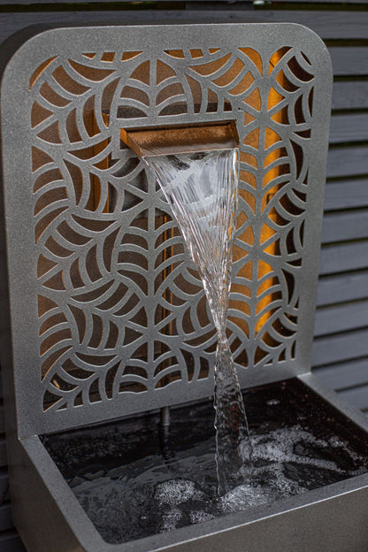 The RENÉ FALLS by La Hacienda is a decorative, self-contained water fountain that features an intricate geometric pattern. Water elegantly cascades from a rectangular spout into the basin below, set against horizontal grey slats and enhanced by warm lighting for an art deco ambiance.