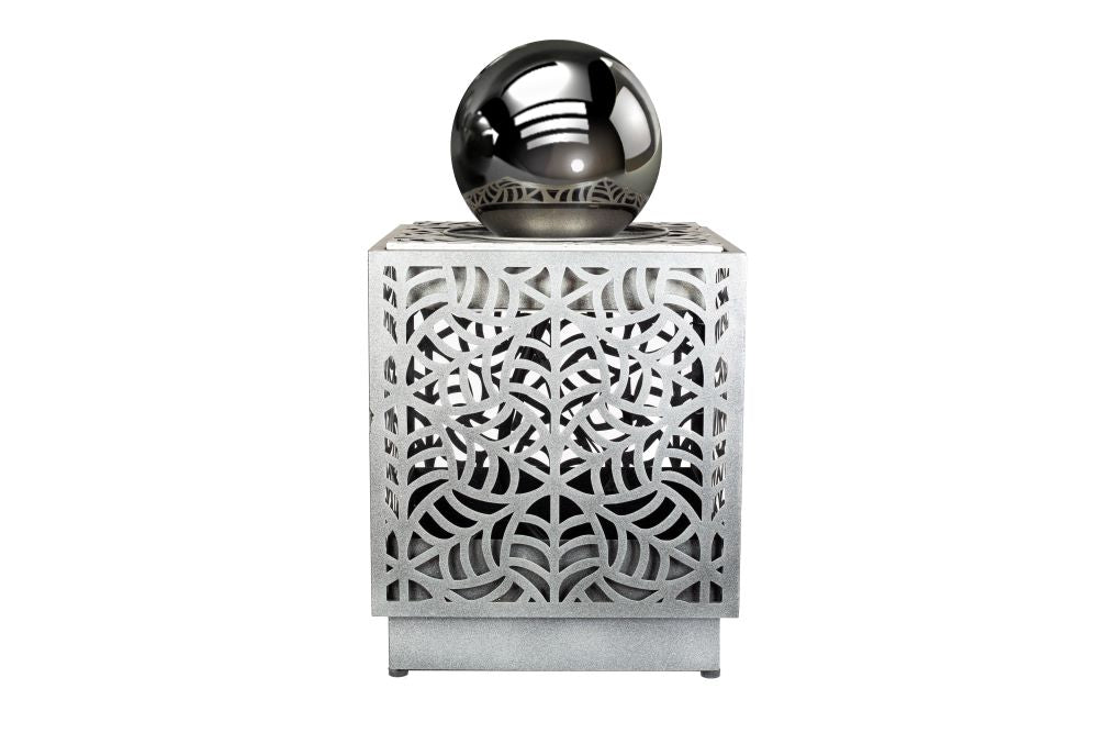 The RENÉ CASCADE by La Hacienda is a decorative cube adorned with intricate cut-out patterns and crowned by a reflective metallic sphere on a white base, evoking the appearance of a self-contained fountain. Its design incorporates geometric and curvilinear motifs, offering a modern artistic aesthetic.