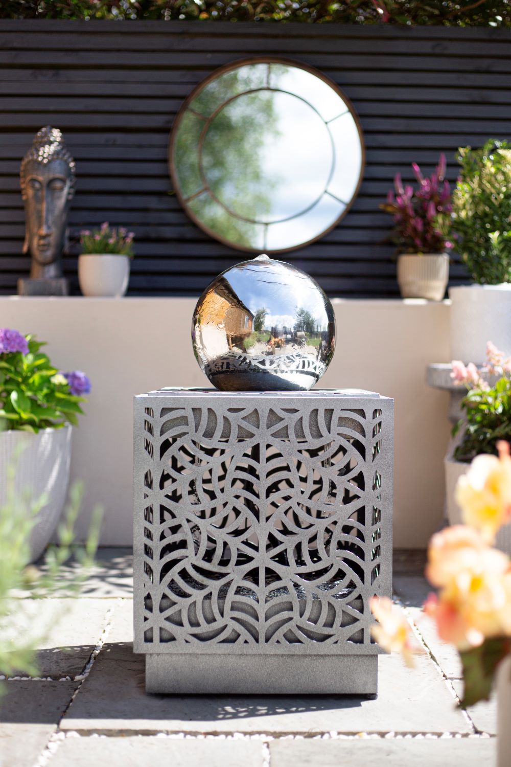 A tranquil outdoor space featuring the La Hacienda Rene Cascade Water Feature, which includes a decorative stone cube with intricate patterns topped with a reflective silver sphere and incorporating an elegant water feature powered by a recirculating pump. In the background, potted plants, a bust sculpture, and a circular wall mirror reflect the lush greenery and vibrant flowers.