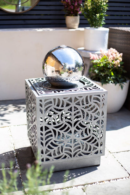 A RENÉ CASCADE by La Hacienda, featuring a sleek sphere perched on an intricately patterned cube that mimics the look of a self-contained fountain, is displayed on a stone patio. In the background, there are potted plants and a wall adorned with horizontal wooden slats.