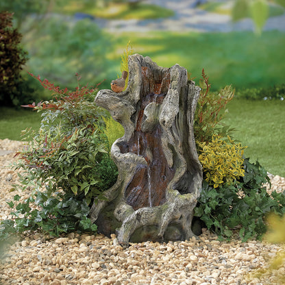 A decorative garden scene showcases a faux tree stump water feature, accentuated by La Hacienda's Willow Spills inc LEDS, surrounded by lush green and yellow plants. It's set atop a bed of small pebbles with a small bird perched on the stump. The tranquil background features a softly blurred grassy landscape.