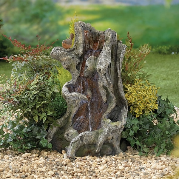 A garden scene featuring a decorative sculpture of a twisted tree stump with a small animal perched on it is beautifully enhanced by La Hacienda's Willow Spills inc LEDS. The surrounding green foliage and plants, set on a pebbled surface with the LED lights providing a gentle glow, create an enchanting atmosphere.