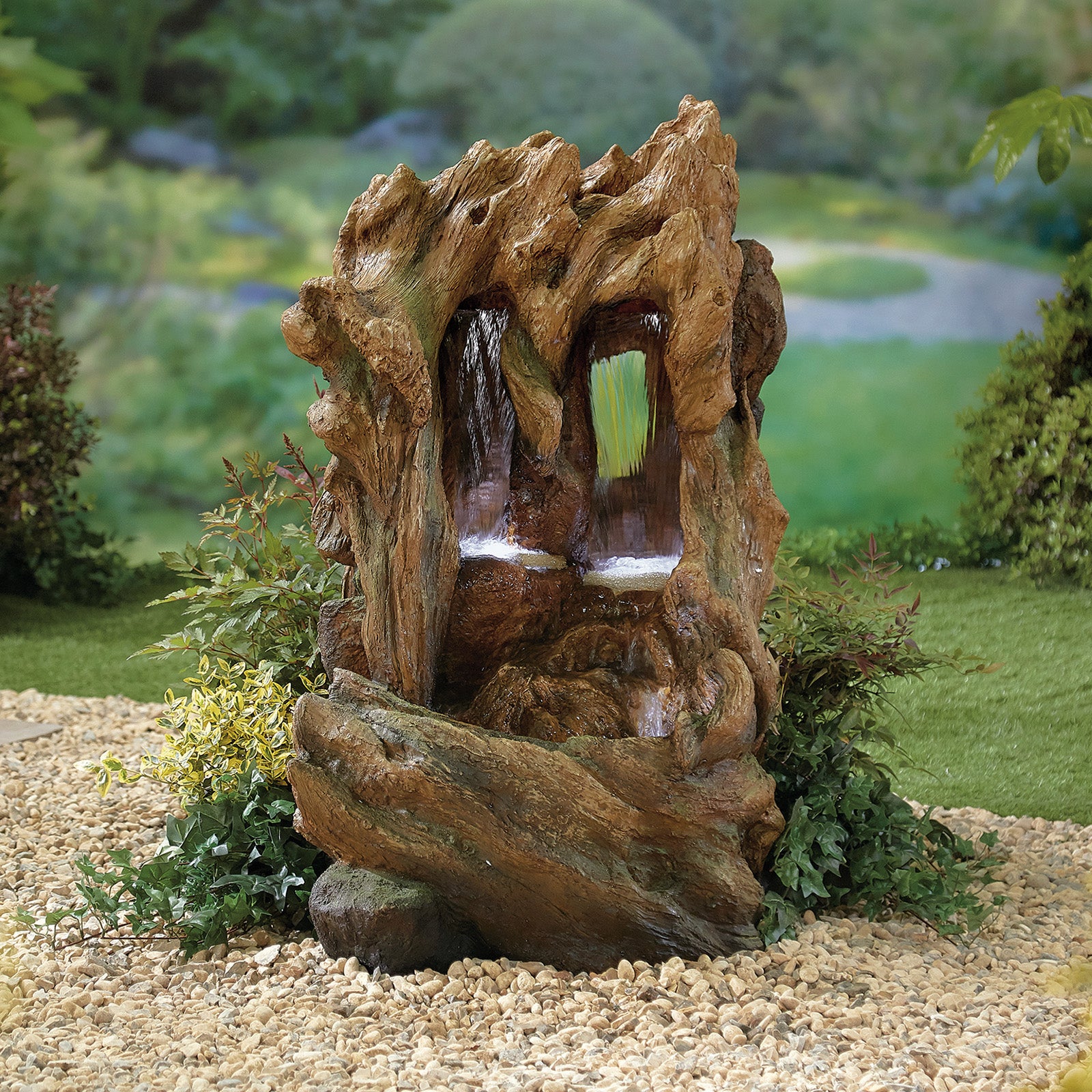 La Hacienda's Colorado Falls inc LEDS is a garden water feature designed to resemble a rustic tree trunk, crafted from hand-painted resin. The water flows through two openings and is surrounded by small green plants and gravel, with a blurred garden landscape as the background.