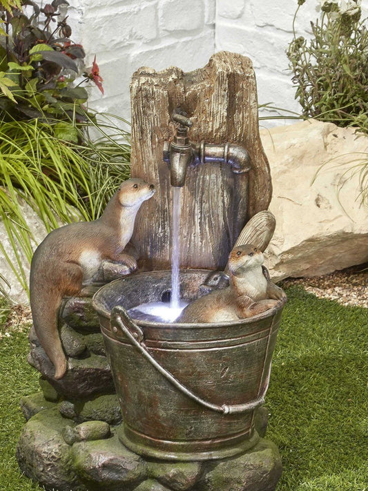 The Otter Pools inc LEDS by La Hacienda is a decorative garden fountain featuring two otter sculptures. One otter leans on the edge of a metal bucket filled with water, while the other peeks over the rim. The setup is enhanced by LED lights and includes a wooden post with an attached faucet for a self-contained design.