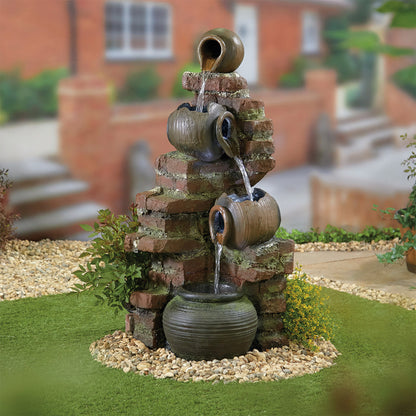 The La Hacienda Flowing Jugs inc LEDS is a delightful garden fountain where water gracefully cascades from rustic clay jugs onto a brick structure. This charming feature is enhanced with LED lights and flows into a round basin nestled on a gravel bed. Surrounded by grass and plants, it serves as perfect garden decor against the softly blurred building in the background.