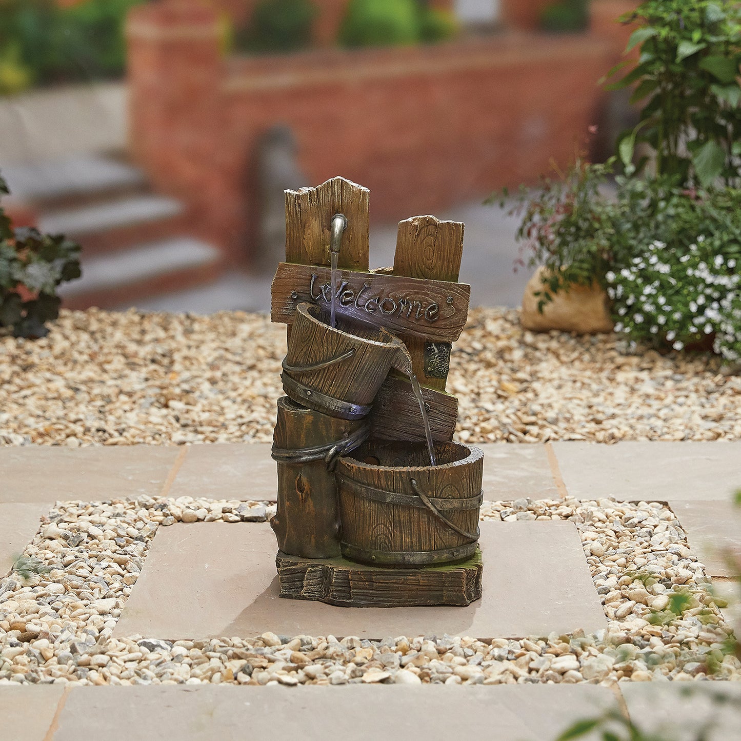 Transform your garden with a delightful decorative scene featuring the Fence Post Pours with LEDS by La Hacienda. This rustic wooden fountain includes a welcoming "Welcome" sign and elegantly directs water through a small pipe into barrels, enhancing its water feature charm. It is beautifully set among pebble stones and greenery, complete with the inviting backdrop of steps and brick walls.