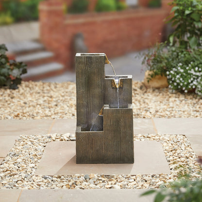 A Coastal Sleepers inc LEDs water feature from La Hacienda elegantly enhances a pebbled area with its cascading design across three levels. The installation is beautifully framed by a paved square and lush green plants, with blurred stone steps providing a picturesque backdrop.