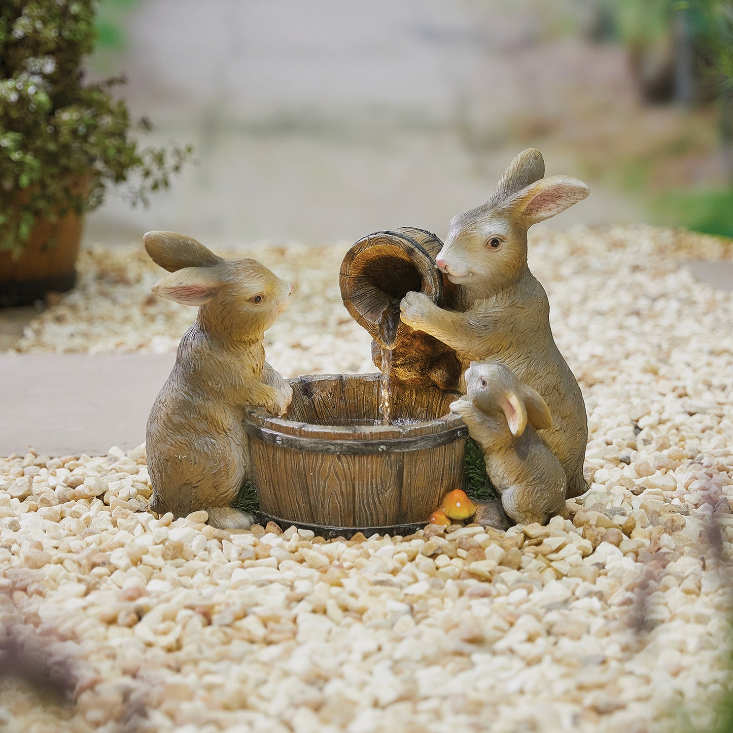 Three charming bunny sculptures from La Hacienda's "Playful Bunnies inc LEDs" are situated around a small barrel fountain on a gravel surface. One rabbit is depicted pouring water from a jug into the barrel as the others look on. Delicate LED lights enhance the scene, while plants and greenery softly blur in the background, adding to this enchanting water feature.