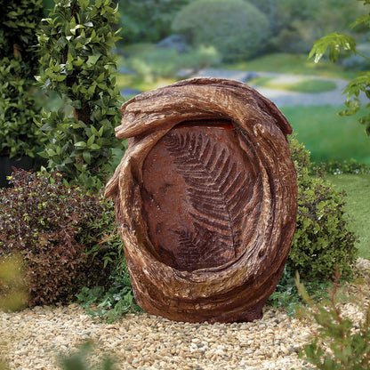 In a picturesque garden setting, the Forest Fern inc LEDs by La Hacienda is showcased as an impressive large decorative sculpture inspired by a fossilized fern encased in twisted wood, elegantly functioning as an LED-illuminated fountain. It is beautifully nestled among lush greenery and small stones, with a softly blurred garden landscape in the background.