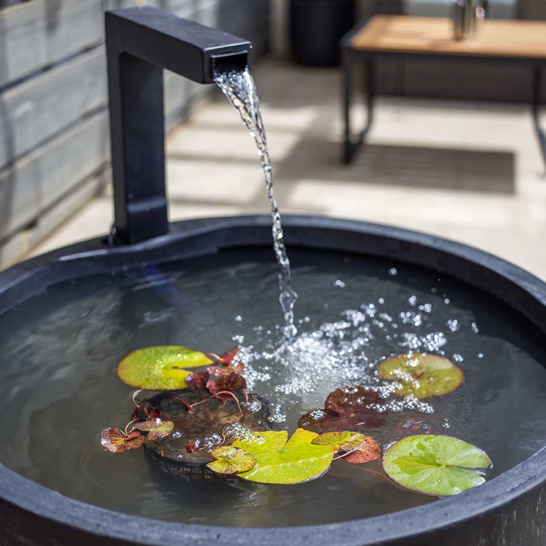 An upgraded outdoor space featuring the La Hacienda Patio Pond includes a contemporary fountain where water elegantly cascades from a square black spout into a circular basin. The basin is beautifully decorated with floating lily pads and small aquatic plants, enhancing sunlit areas where a table can also be seen.