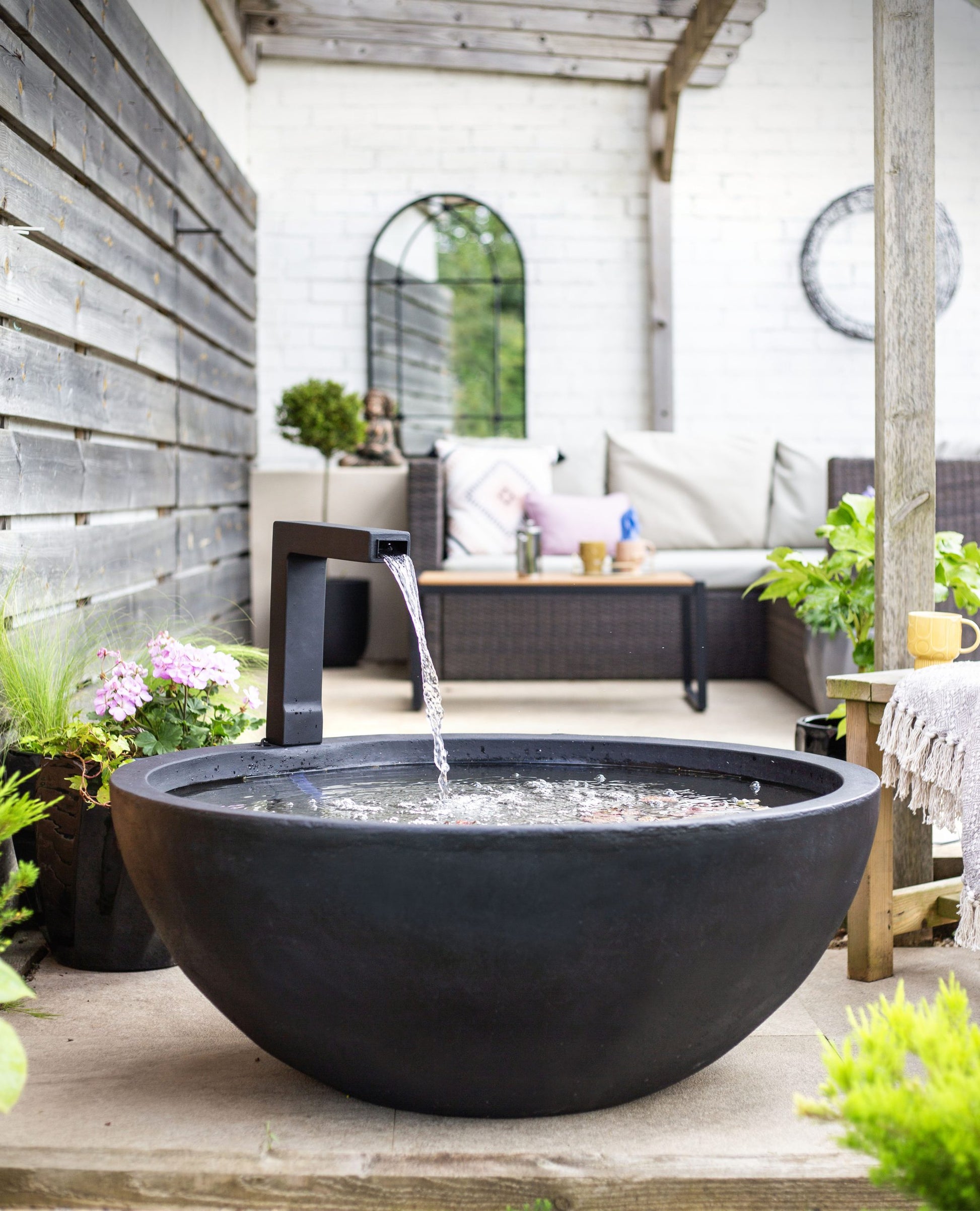 A serene outdoor space showcases the Patio Pond by La Hacienda, a black stone water fountain that adds a calming water feature to the foreground. A wicker sofa with beige cushions is nestled under a pergola, surrounded by lush plants. The peaceful ambiance of this exquisite patio retreat is further enhanced by a round mirror and wooden wall.