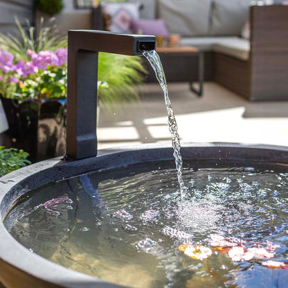 A modern Patio Pond by La Hacienda, featuring a sleek black water fountain, pours a gentle stream into its circular basin, creating a tranquil setting. Floating petals enhance the serene surface while outdoor spaces in the blurred background, equipped with furniture and purple flowers, add to the cozy atmosphere.