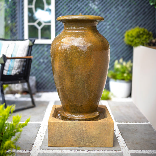 A tall Athenian Vase by La Hacienda, reminiscent of Grecian vases, sits on a stone base. It's surrounded by green plants and a modern patio setting with a cushioned chair and geometric wall patterns in the background.