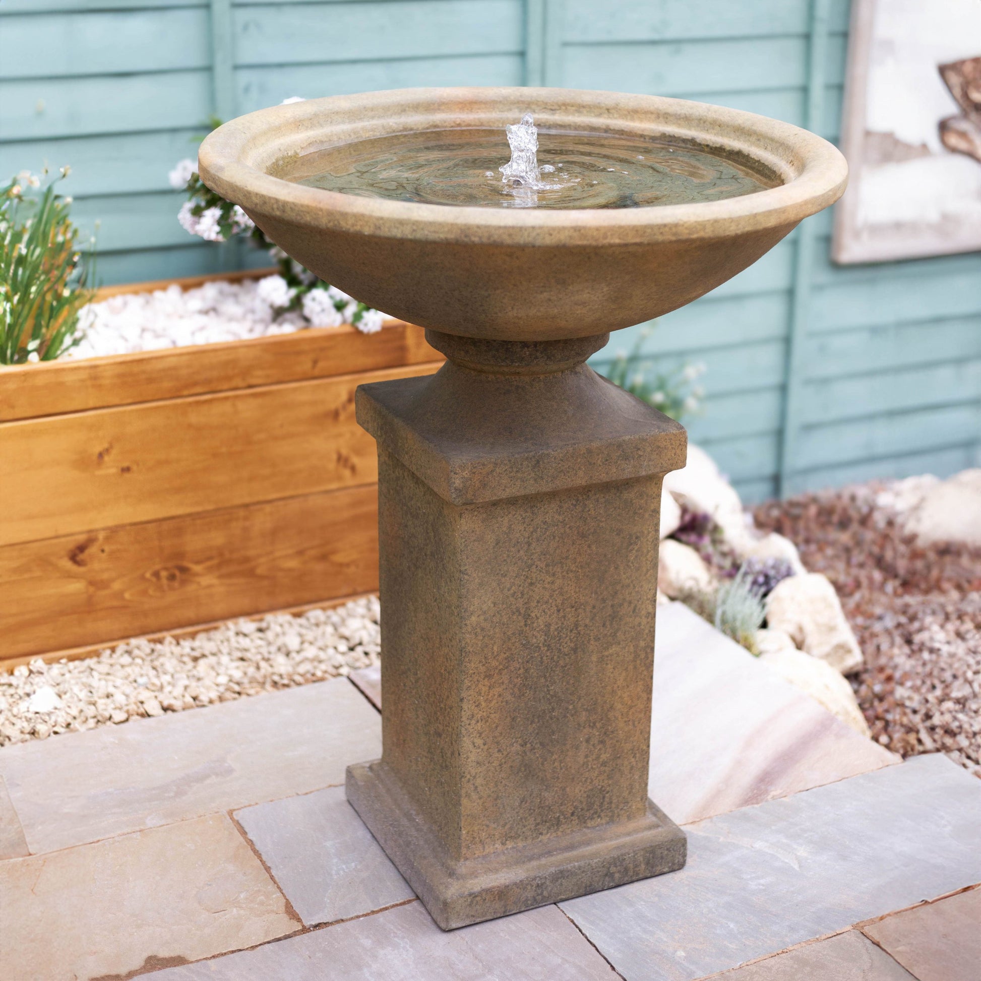 A Roman Pedestal from La Hacienda is elegantly placed on a paved patio, featuring a wide basin that gently bubbles water at the center, adding charm to the garden. Surrounding it is a wooden planter filled with plants, a gravel area, and a blue painted fence in the background.