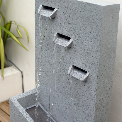 The "Trinity Falls incl LEDs" by La Hacienda, a sleek gray stone fountain with three elegantly cascading spouts into a rectangular basin, showcases contemporary garden decor. Complementing the scene is a vibrant green plant in a white planter, bringing nature's charm as it sits on a wooden floor against a pale wall.