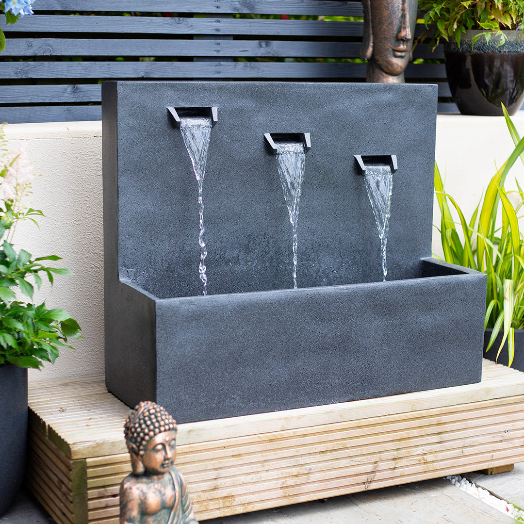 A rectangular stone fountain from La Hacienda, known as the Kabe Falls incl LEDs, features three spouts cascading water into a basin and is elegantly positioned on a wooden platform to create a serene focal point in your garden. This outdoor feature is beautifully surrounded by lush green plants and a Buddha statue, with the ensemble complemented by an artistic decorative screen and another statue in the background.