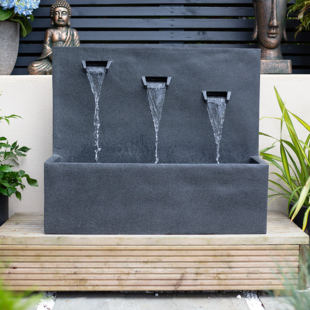 The Kabe Falls incl LEDs by La Hacienda is a contemporary outdoor water fountain featuring three spouts that cascade water into a rectangular basin. As an elegant centerpiece for your garden, it is beautifully displayed on a wooden platform and surrounded by plants and decorative sculptures, such as a Buddha statue, to create a serene and tranquil outdoor setting.