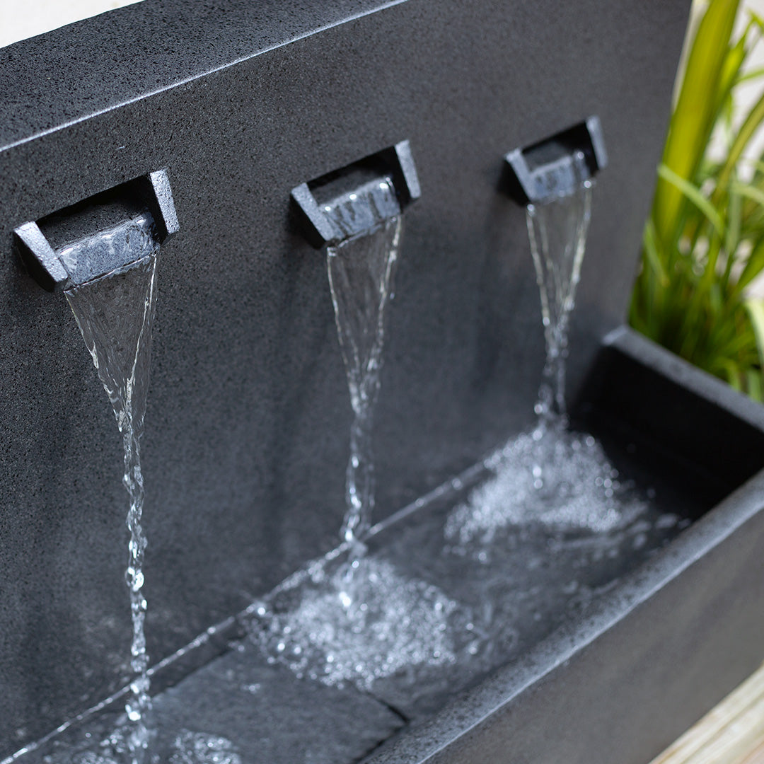 Close-up of the modern Kabe Falls incl LEDs, a three-tiered outdoor water feature by La Hacienda, with water flowing from three spouts into a rectangular basin below. Made of dark stone, this striking garden piece beautifully contrasts against a lush backdrop of green plants.