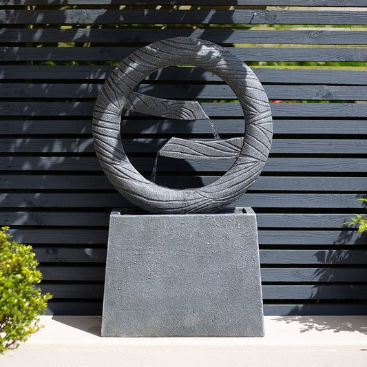 La Hacienda's Jackson Spills, a contemporary stone water feature with a circular design, enhances the garden oasis when placed against a horizontal black slatted fence. Elegant twin streams of water cascade from the circular structure into a square basin surrounded by lush greenery.