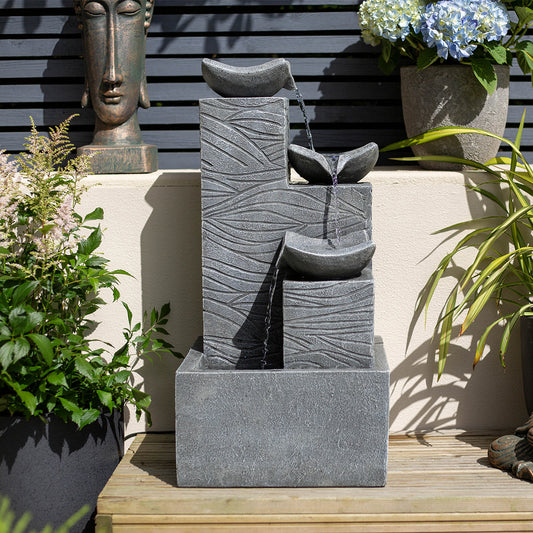 A stylish, tiered stone water fountain with a modern geometric design flows gently, embodying the essence of contemporary garden water features. Surrounded by potted plants and a Buddha sculpture in the background, this La Hacienda Jackson Pours incl LEDs creation radiates serenity and invites relaxation.