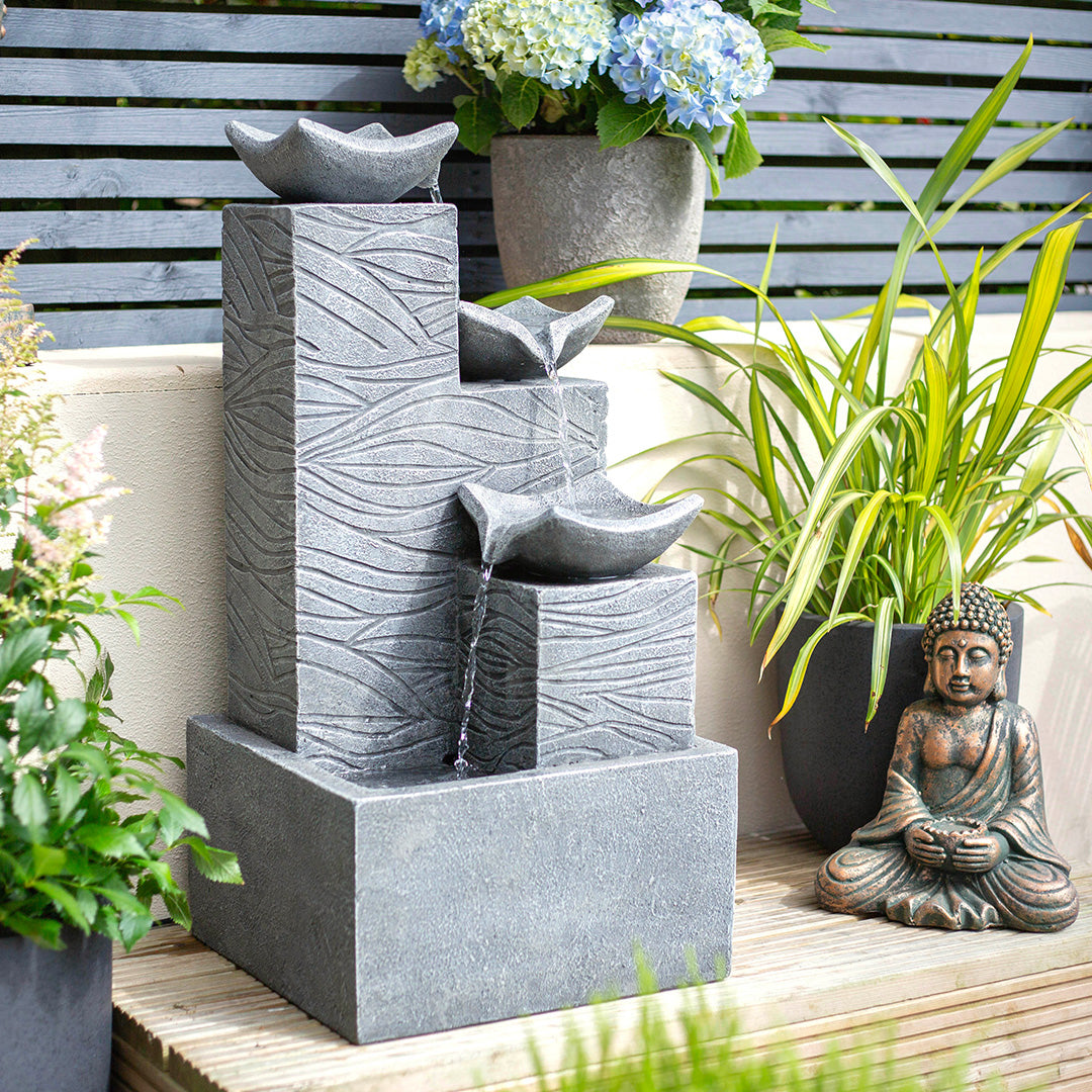 A modern gray stone water fountain, known as Jackson Pours incl LEDs from La Hacienda, stands out among garden water features with its tiered design, where water cascades through leaf-shaped bowls. Nearby, surrounded by potted hydrangeas and grasses, a small Buddha statue meditates peacefully on a wooden surface.