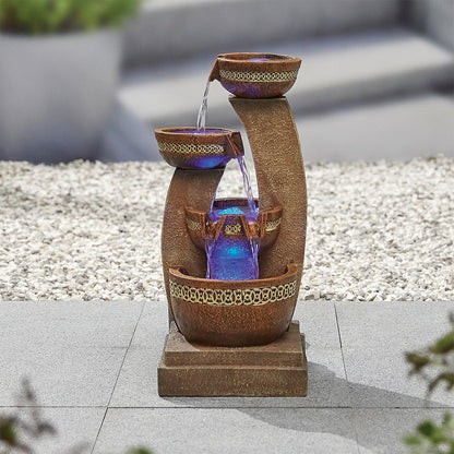 The Azure Columns inc LEDS by La Hacienda is a poly-resin, stone-like tiered garden fountain that boasts three levels of bowls. Water elegantly cascades down each tier, accompanied by the soothing blue glow of LED lights in the middle section. This azure water feature stands gracefully on a paved surface against a gravel backdrop.