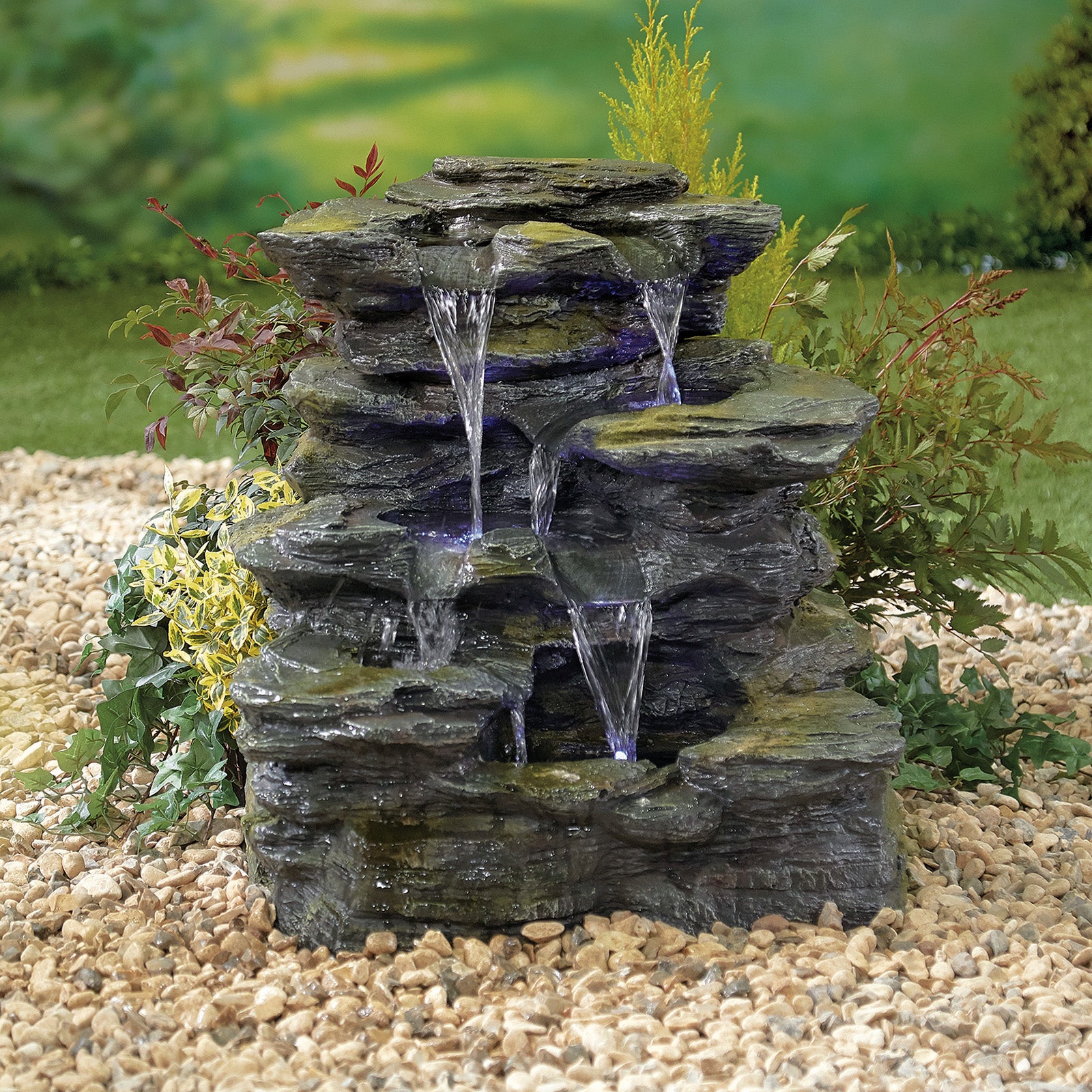 The Como Springs inc LEDS by La Hacienda is a cascading rock fountain where water gently flows over multiple tiers, surrounded by small pebbles and lush green plants, set against a blurred natural background. This elegant natural stone water feature enhances any space with its tranquil charm.