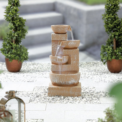 The Sparkling Bowls fountain by La Hacienda, made from poly-resin with cascading water tiers, elegantly enhances the pebble-covered courtyard. Flanked by two manicured potted shrubs, this stunning water feature is illuminated by subtle LED lights that add charm to the steps and greenery visible in the blurred background.