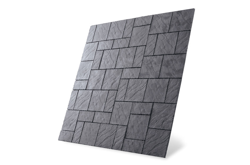 Image of a large rectangular panel featuring the Welsh Slate Chalice Paving Kit by Bowland Stone from the Brisks brand, showcasing a pattern of interlocking rectangular tiles in shades of dark gray. The tiles have a textured, rough appearance, and the panel is set against a plain white background. The panel is slightly tilted, adding an interesting element to garden features.