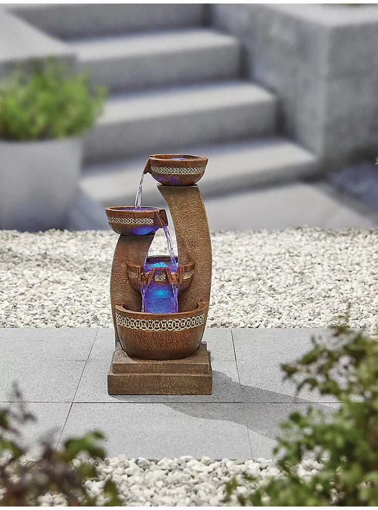 A decorative outdoor fountain crafted with a rustic poly-resin design is beautifully placed on a gravel area. Water gracefully cascades from the top vessel into two lower ones, enhanced by blue LED lights illuminating the base. Stone steps and potted plants in the blurred background further elevate the charm of this Azure Columns inc LEDS water feature by La Hacienda.