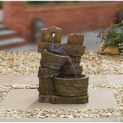 A rustic wooden garden fountain, the Fence Post Pours inc LEDS by La Hacienda, is a delightful addition to your garden decor ensemble. It features a "Welcome" sign with water gently cascading into a barrel. Positioned on a pebble and stone surface, it beautifully enhances the blurred greenery and stairs in the background.