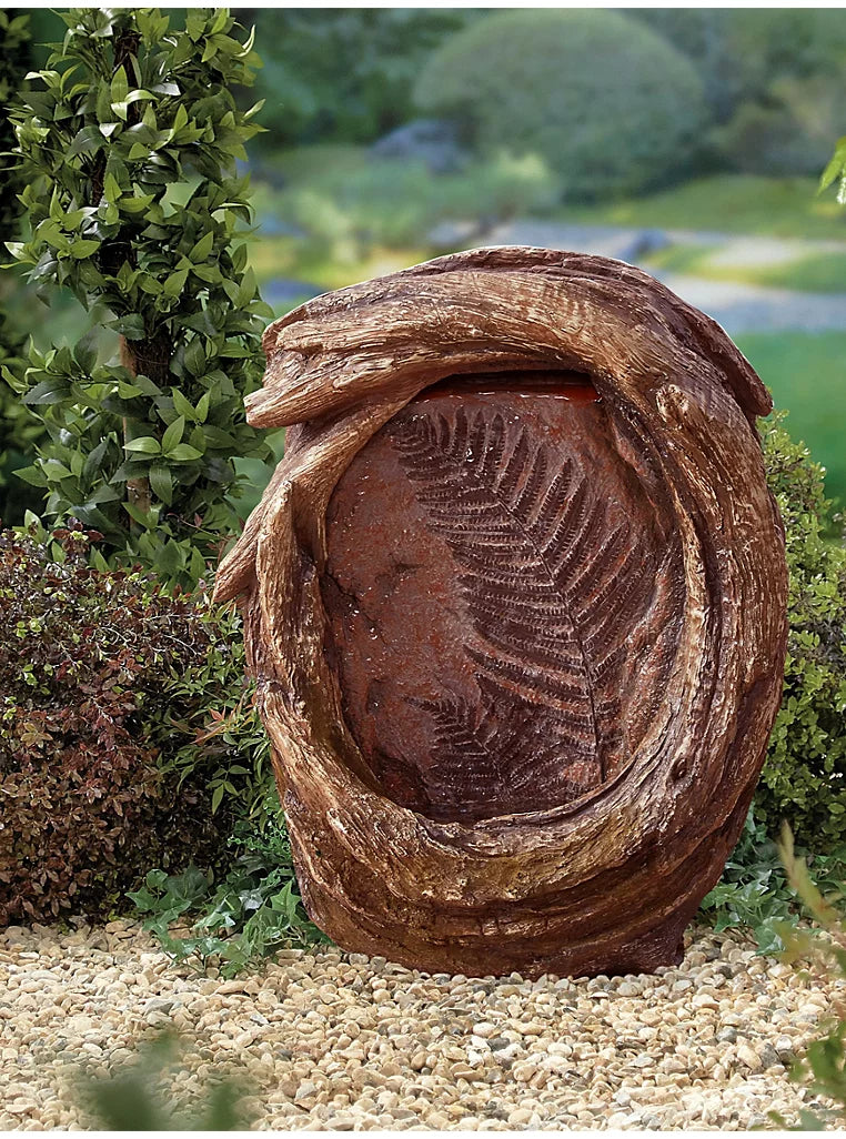 The Forest Fern inc LEDs by La Hacienda is a decorative garden fountain resembling a tree trunk with a carved fern design on its smooth, reddish-brown surface. This resin water feature is complemented by green foliage and plants, with a gravel path in the foreground, creating an enchanting and natural forest vibe.