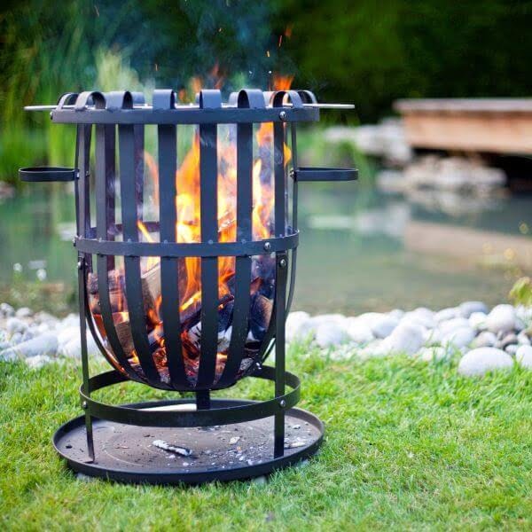 A stylish La Hacienda Vancouver Firebasket is placed on a grassy area, with logs burning brightly, making it ideal for outdoor cooking. In the background, there's a small pond bordered by rocks and a wooden bridge arches over the water, surrounded by lush greenery.